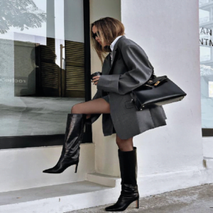 Pessoa de pé ao lado de um prédio branco, olhando pela janela. Veste blazer escuro, shorts e botas pretas altas, carregando uma bolsa preta.